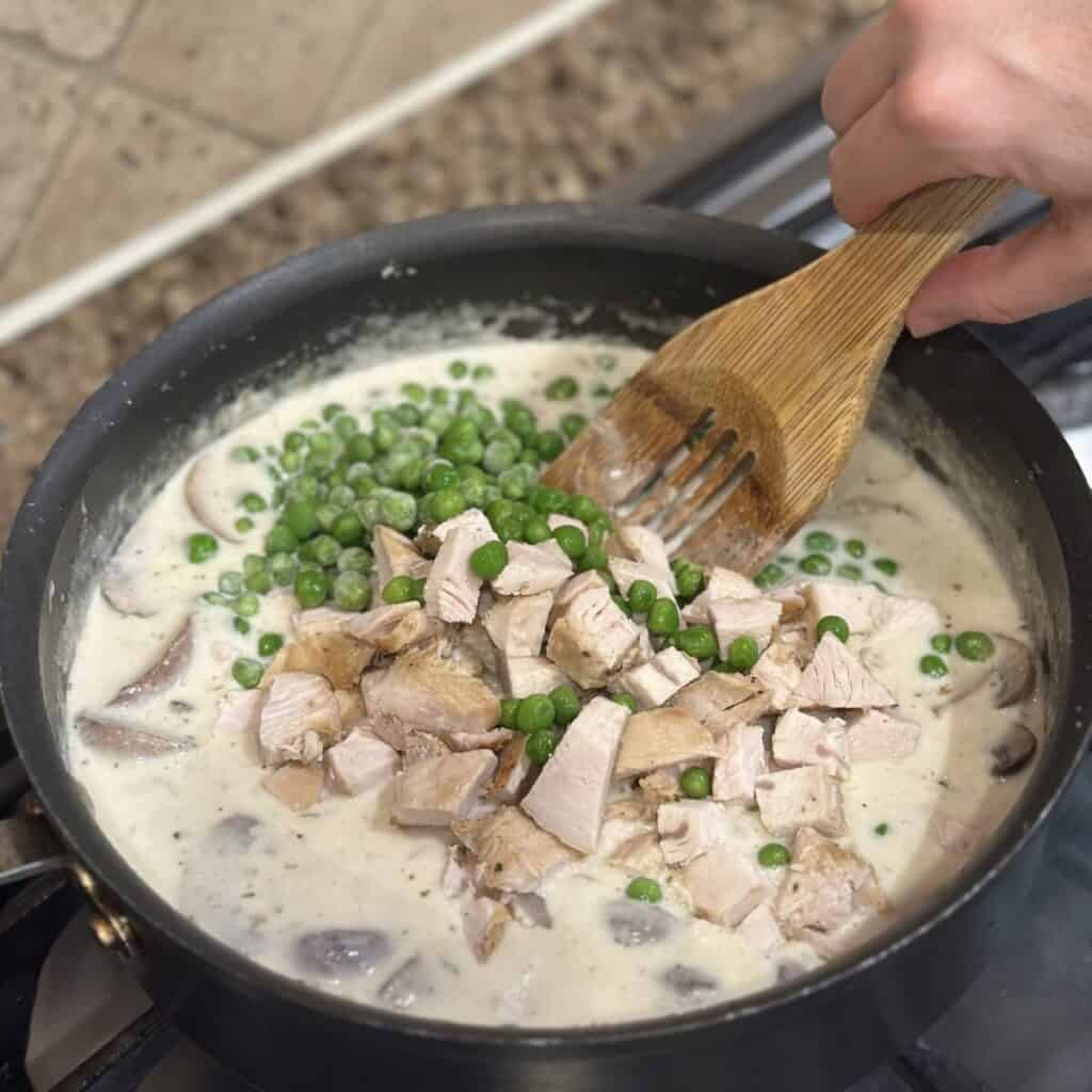 Adding turkey and peas to the filling of turkey tetrazzini in a pan.