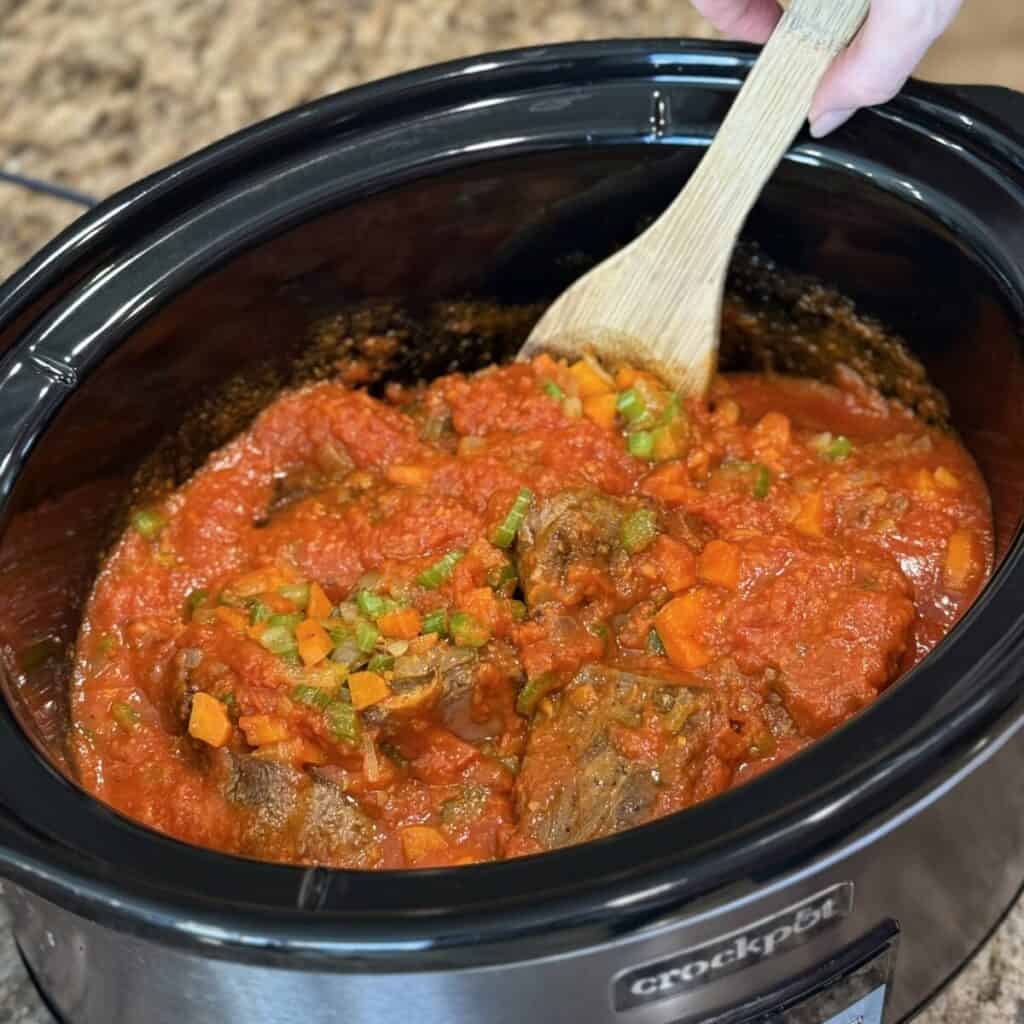 Stirring together marinara, vegetables, and beef ribs in a crockpot.