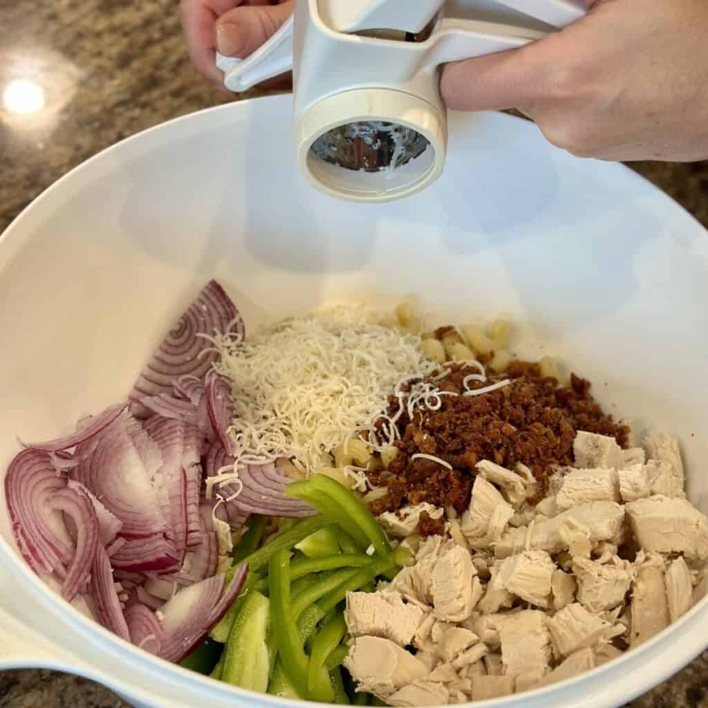 Adding cheese to a bowl filled with ingredients for a pasta salad.