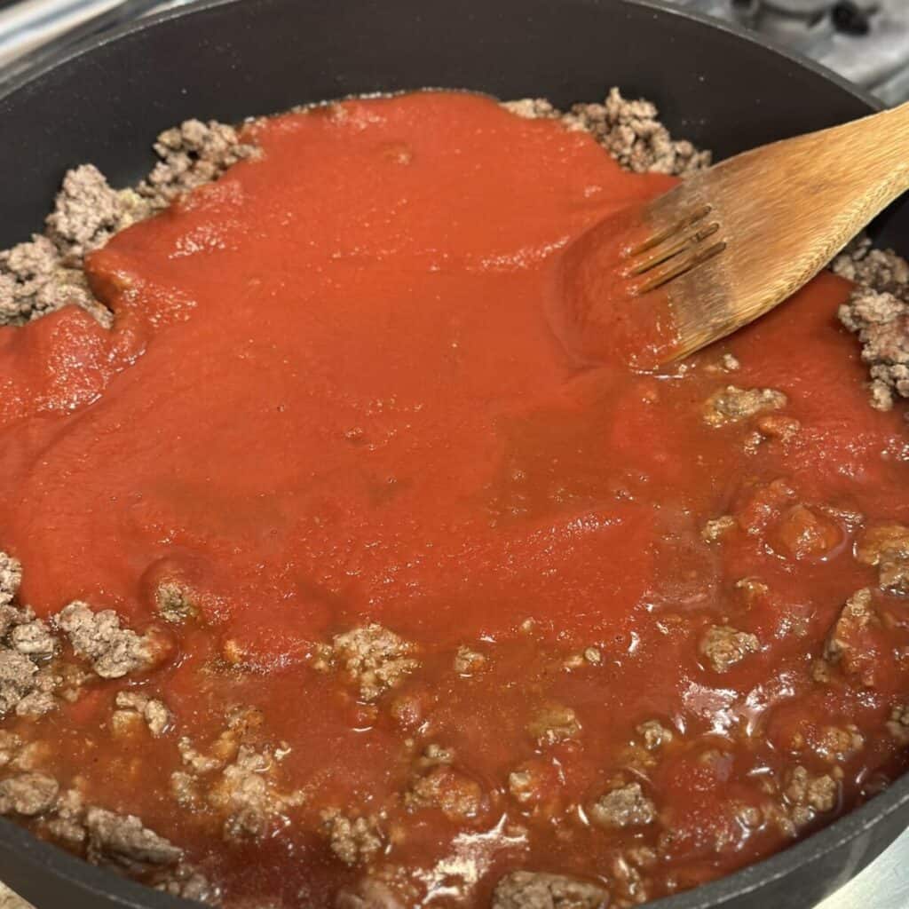 Stirring tomato sauce into ground beef in a skillet.