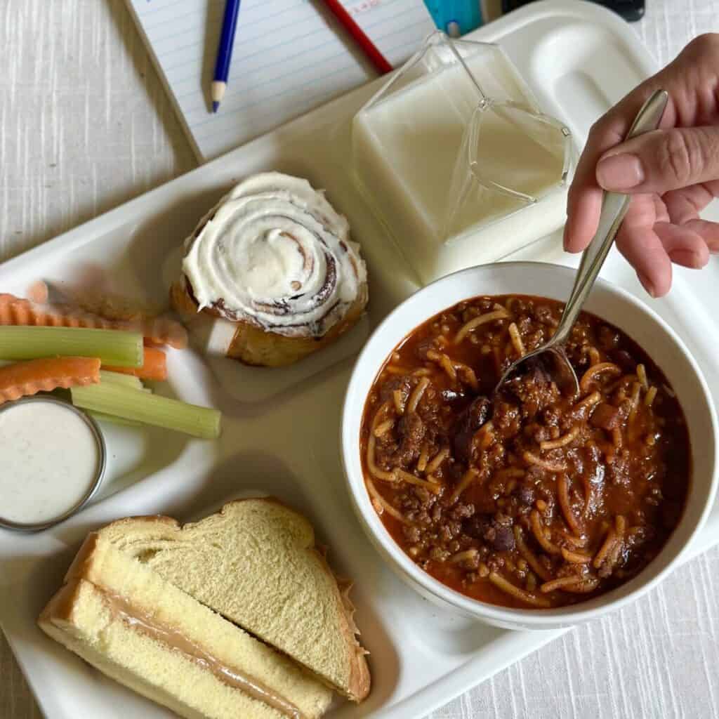 A school tray with chili, veggies, a cinnamon roll, sandwich and milk.