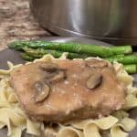 A plate topped with noodles. On top of the noodles is a pork chop with brown gravy. Next to the pork chop is green asparagus.