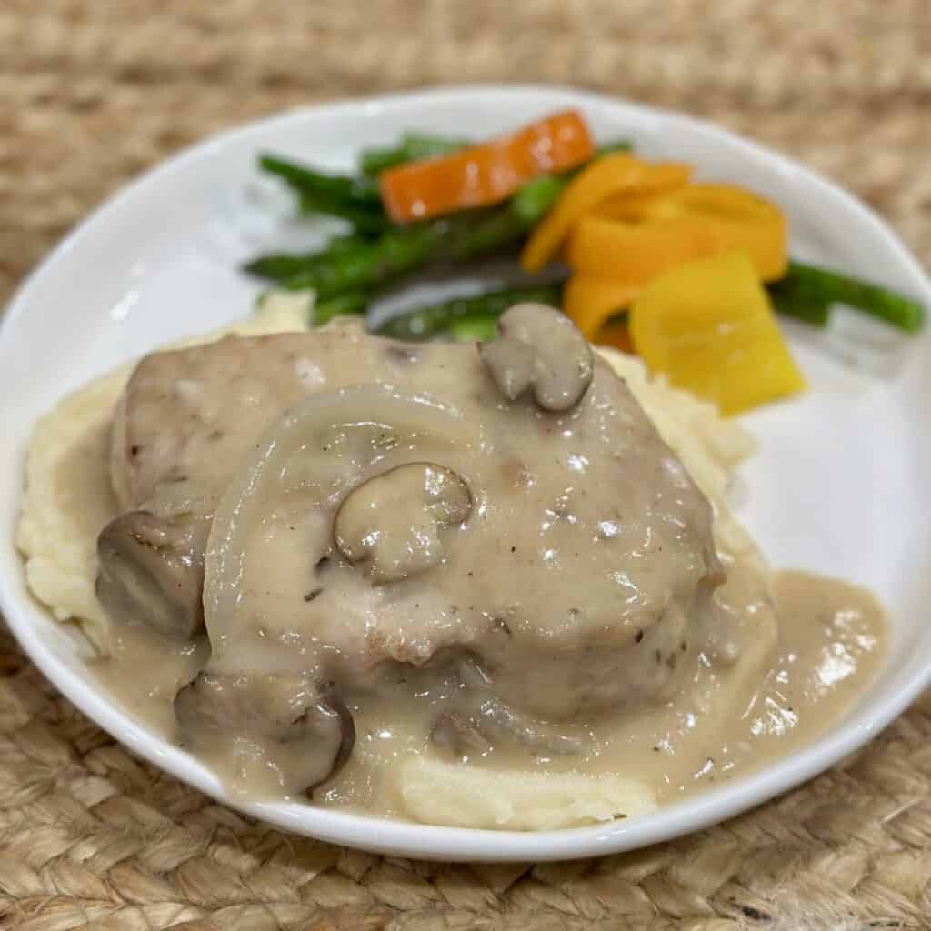 A smothered pork chop over mashed potatoes and vegetables on a plate.