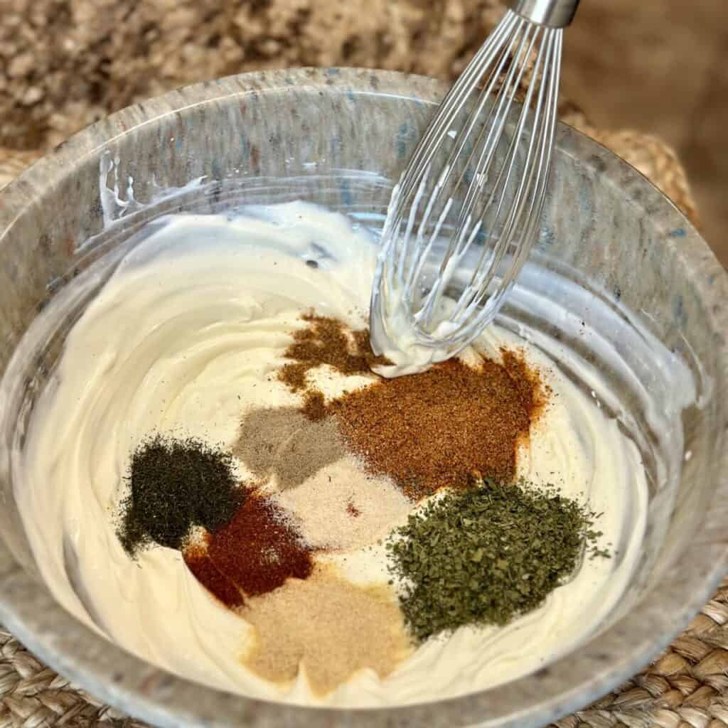 Adding dried herbs to a bowl of mayo, buttermilk and sour cream.