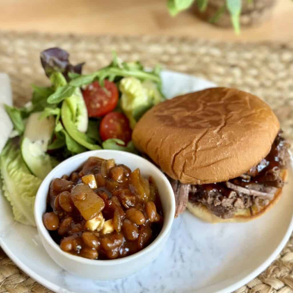 A bowl of baked beans next to a bbq sandwich and salad.