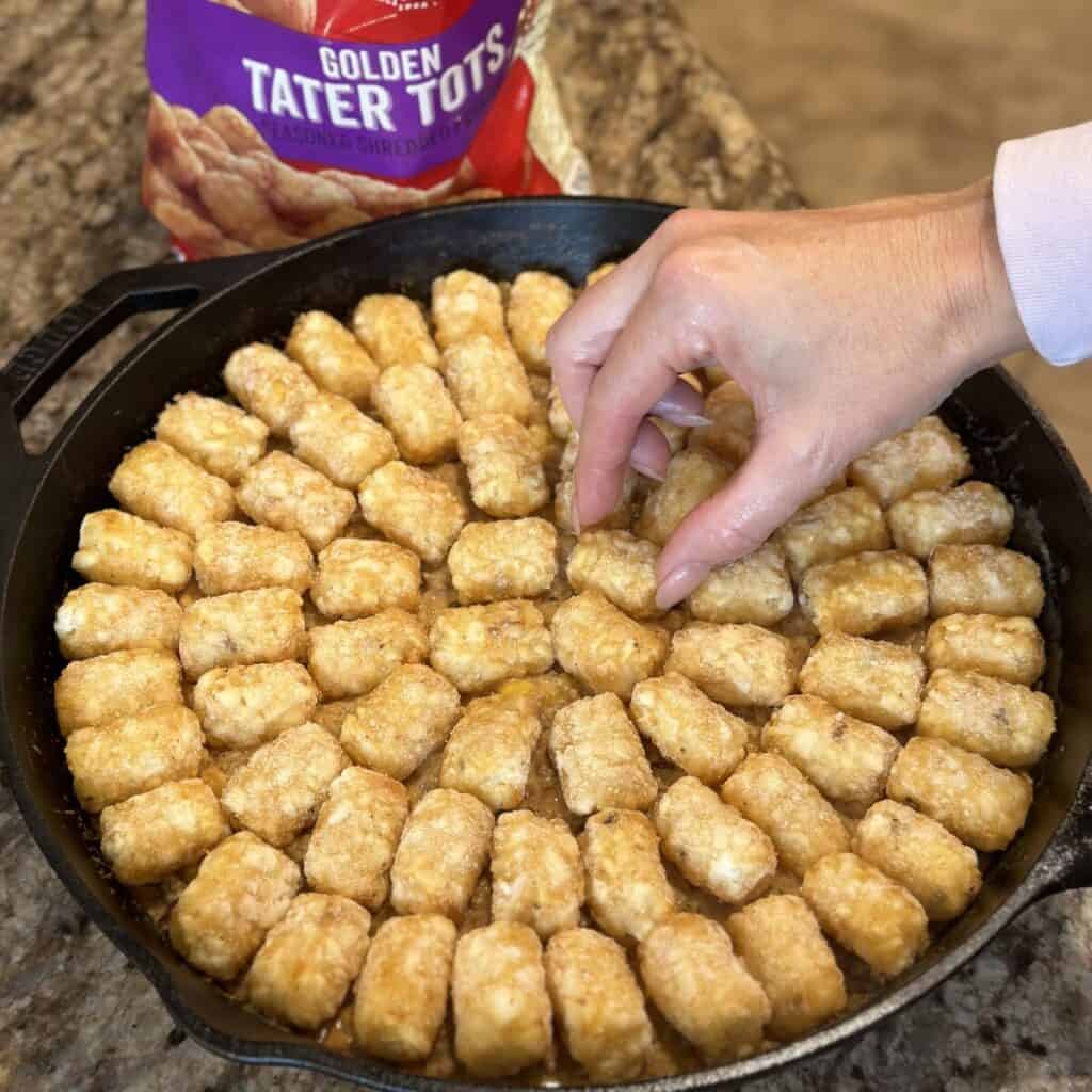 Adding tots to the top of beef in a skillet.