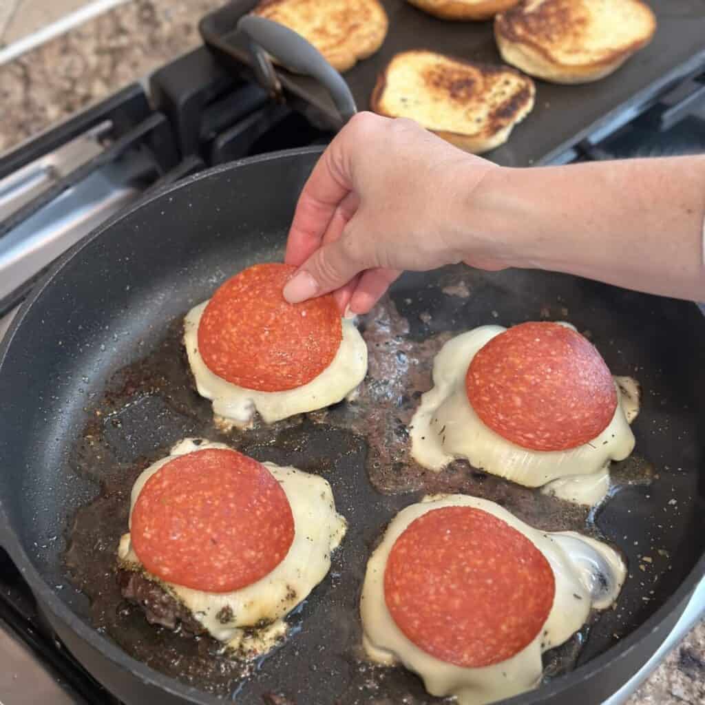 Adding cheese and pepperoni on burgers in a pan.