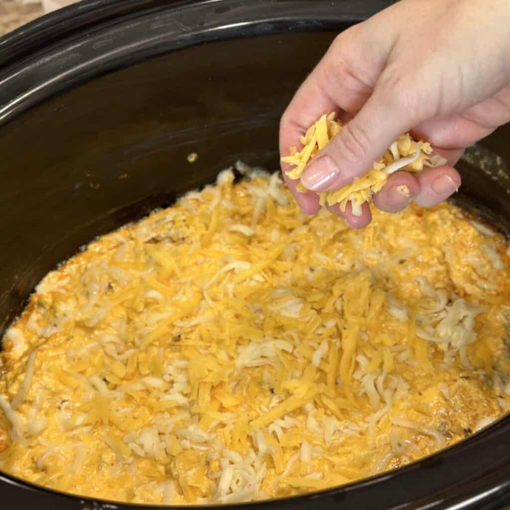 Adding cheese to the top of buffalo chicken dip in a crockpot.