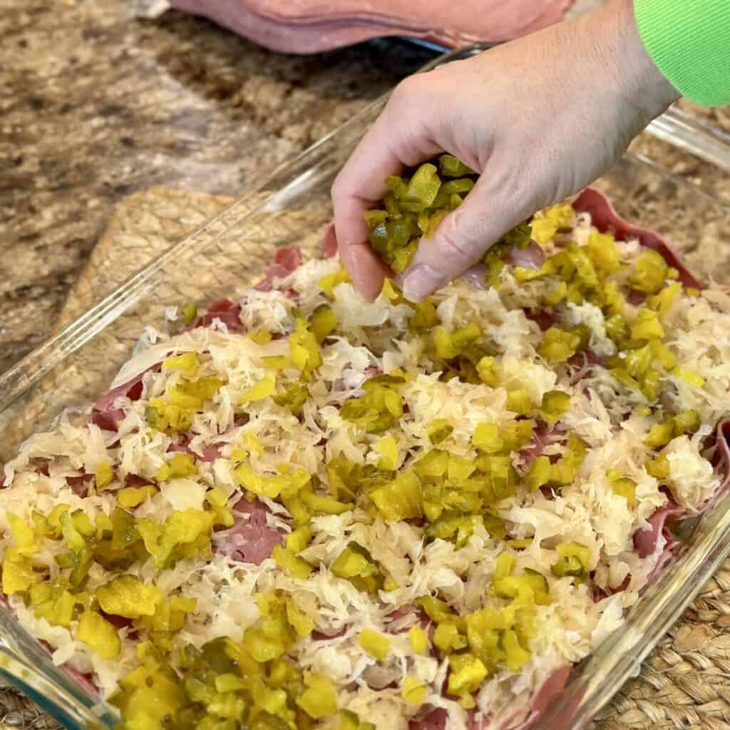 Adding pickles and sauerkraut to the top of a Reuben casserole.