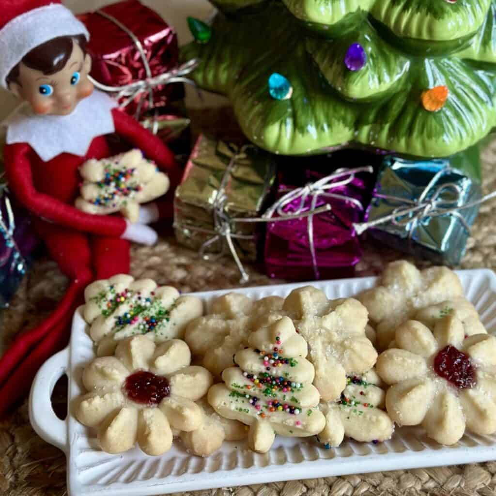 Elf on a shelf holding a half eaten shortbread cookie alongside a plate of cookies.