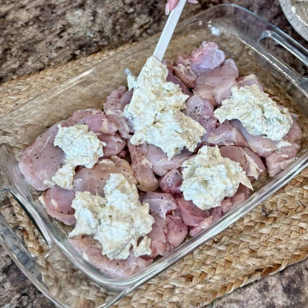 Adding a creamy artichoke spread to the top of chicken in a baking dish.